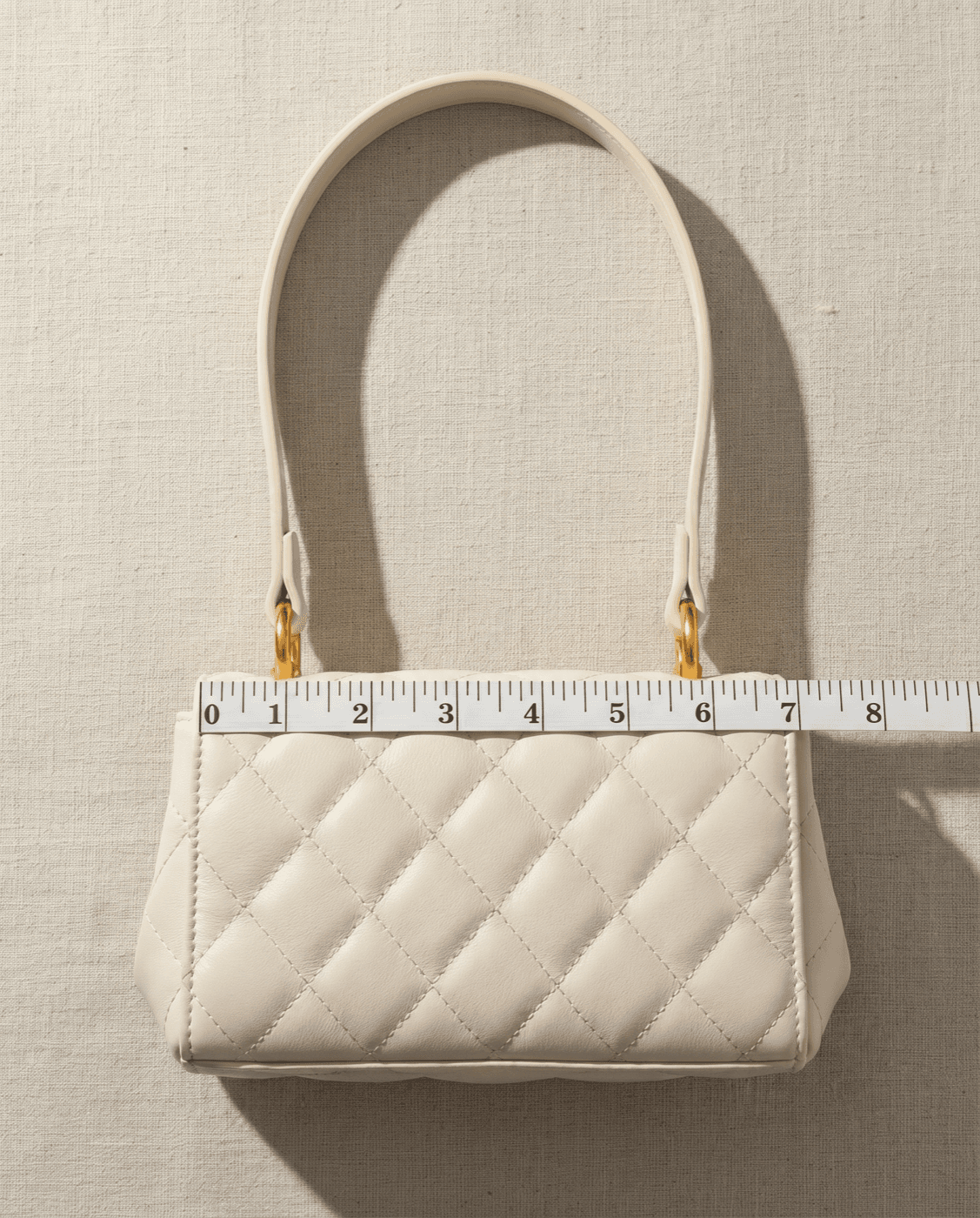 A cream quilted handbag with a white cloth tape measure laid across its top edge to measure the bag's top width for a purse chain length