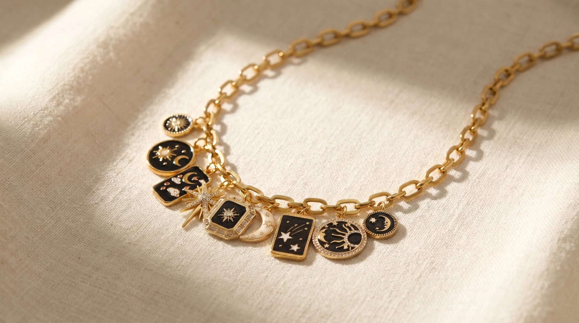 A gold chain charm necklace with celestial-themed black enamel charms on a cream linen surface, by Enleira