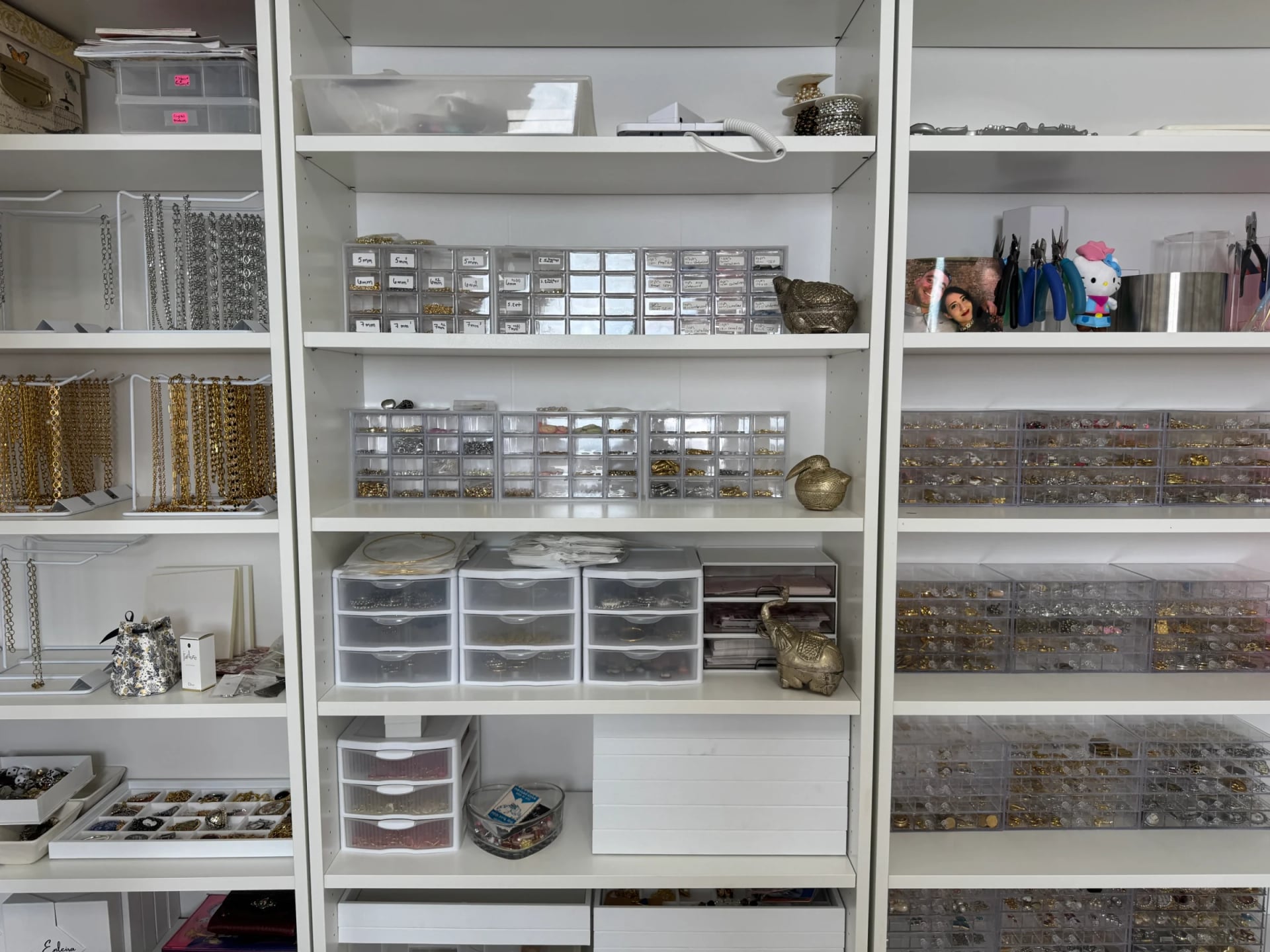 White shelves neatly organized with handcrafted jewelry supplies, charms, and chains for jewelry making, by Enleira