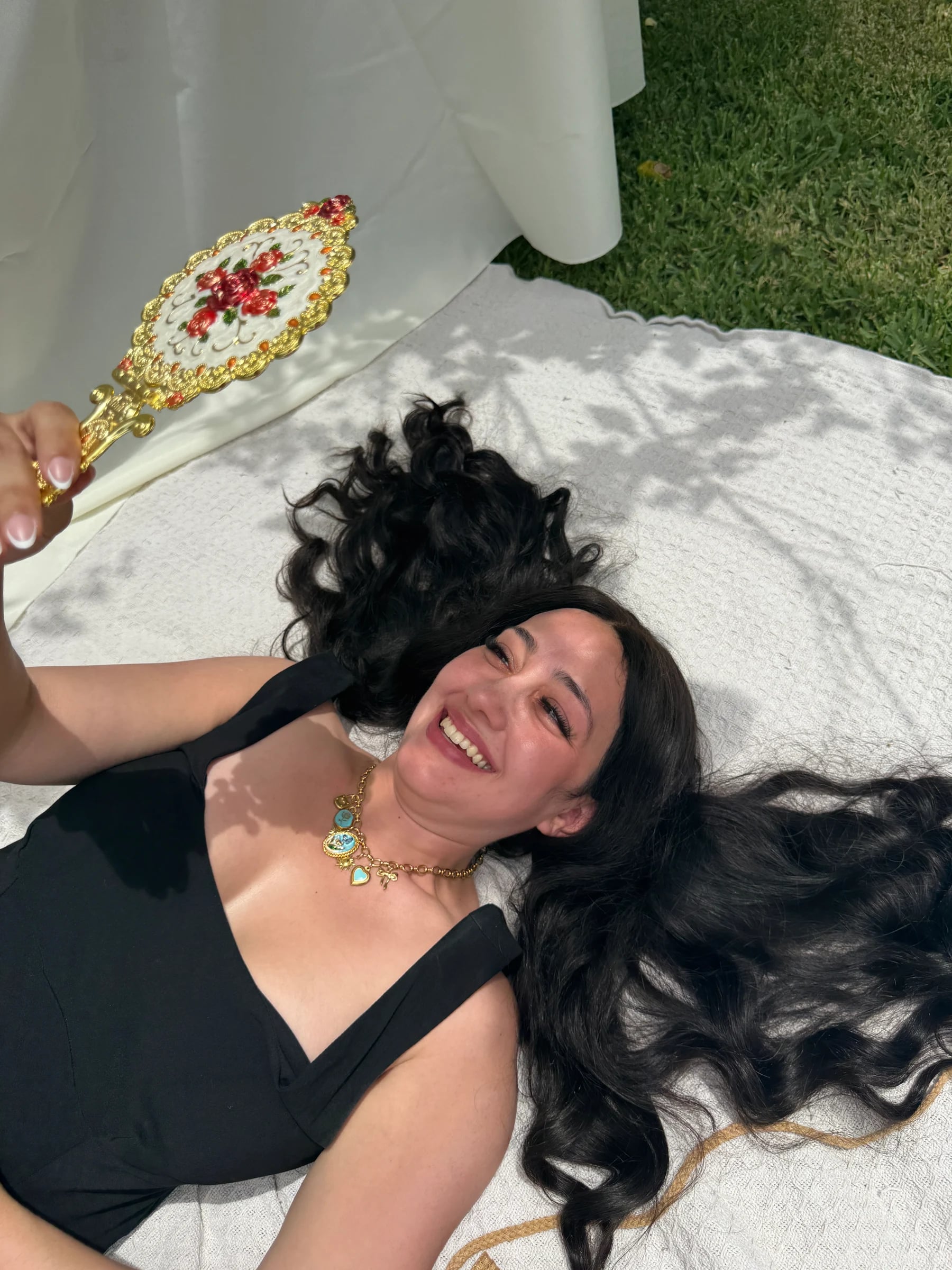 A smiling woman wears a handcrafted 18k gold plated charm necklace while holding a vintage mirror, by Enleira.