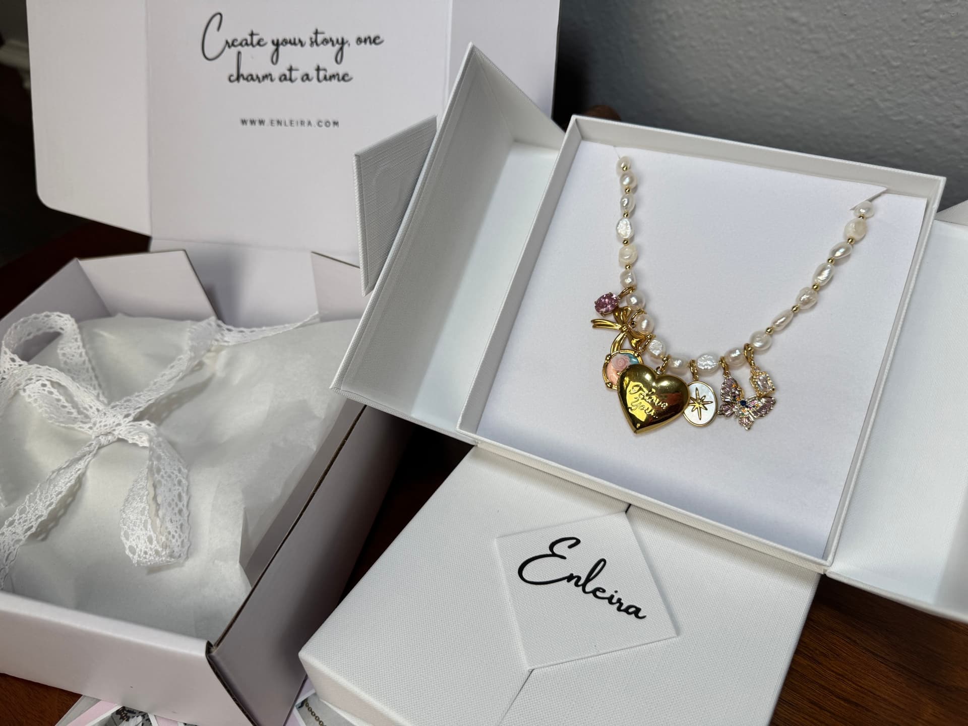 Open Enleira inner jewelry box with a charm necklace on the padded insert, beside the lace-tied tissue parcel and Care Card, by Enleira
