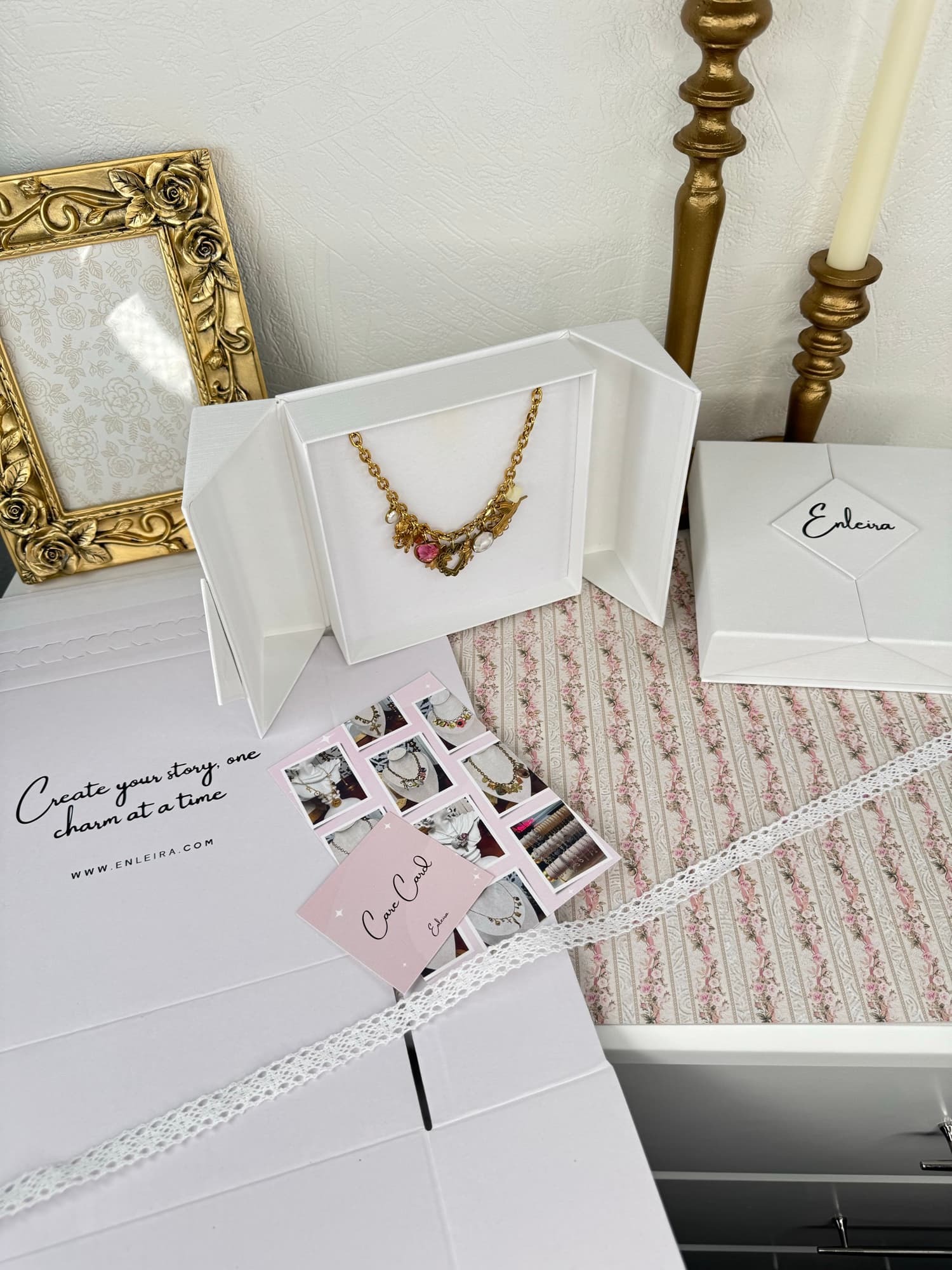 Styled Enleira packaging flatlay — rhombus logo box, blush-pink Care Card, open inner jewelry box, and a charm necklace on linen, by Enleira